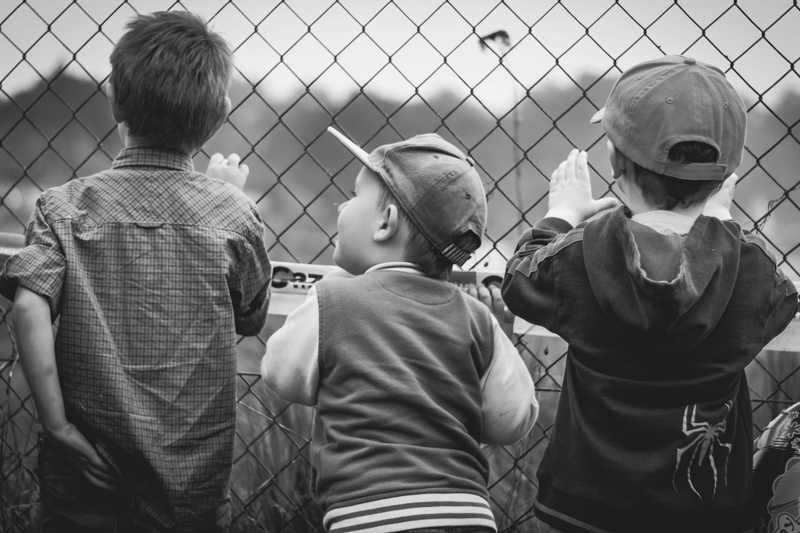 three kids looking through a chain link fence onto a field